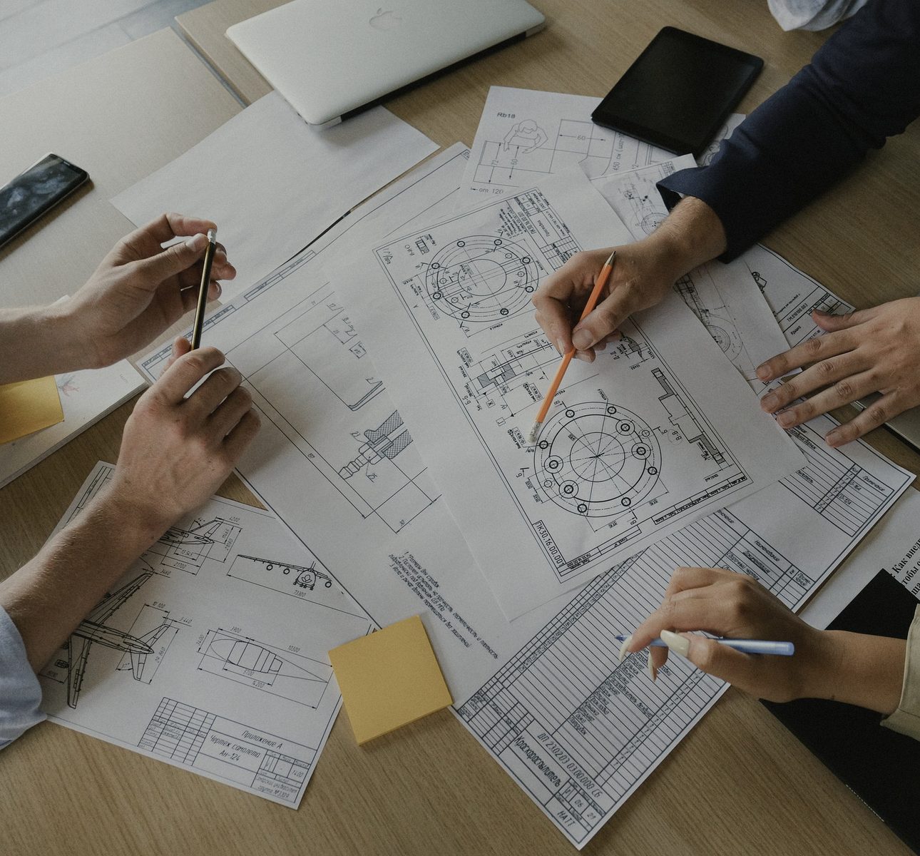 Overhead view of a team working together around one large technical drawing, hands pointing, writing and marking up the shared plan